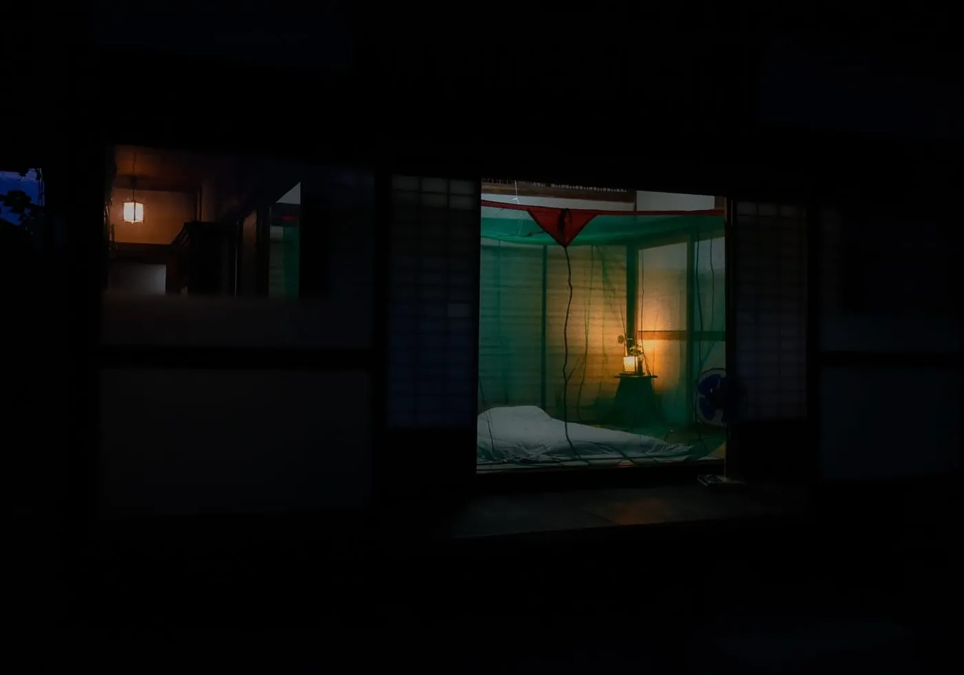 A green mosquito net hung in a tatami room at night, lit by a paper lantern